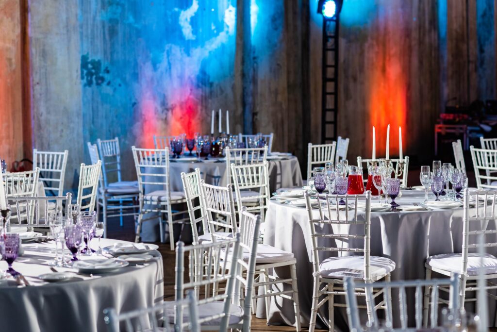Round tables set with linens and centerpieces inside a string-lit wedding reception barn